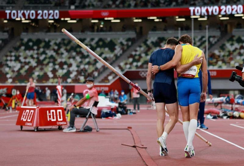 Fotografia em um estádio com o painel “Tokyo 2020”: dois atletas do salto com vara caminham abraçados de costas, um de uniforme azul-escuro e outro de uniforme amarelo e azul.
