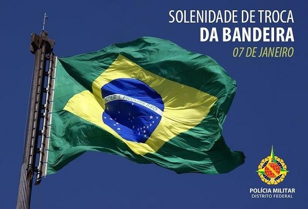 Fotografia da Bandeira Nacional hasteada tremulando em um mastro metálico, com o texto “Solenidade de troca da Bandeira — 07 de janeiro” e o brasão da Polícia Militar do Distrito Federal.