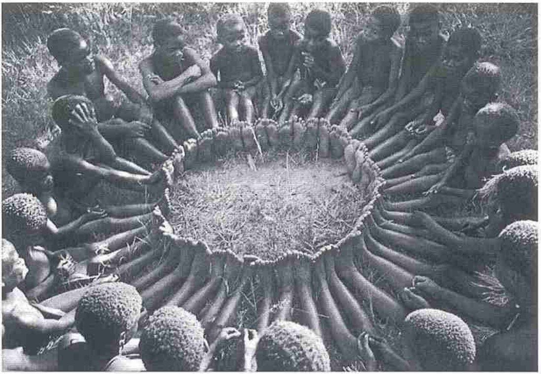 Fotografia em preto e branco: um grupo de crianças africanas sentadas no chão de grama, formando um círculo, com os pés unidos lado a lado no centro, compondo uma roda contínua.