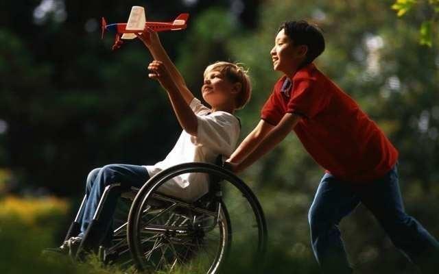 Fotografia de um menino que empurra a cadeira de rodas de outro menino; o menino na cadeira ergue um avião de brinquedo, ao ar livre.