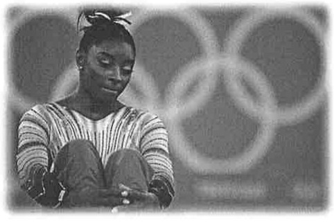 Fotografia em tons de cinza da ginasta Simone Biles sentada, com os joelhos dobrados junto ao corpo e as mãos sobre os pés, olhando para baixo; ao fundo, os anéis olímpicos desfocados.
