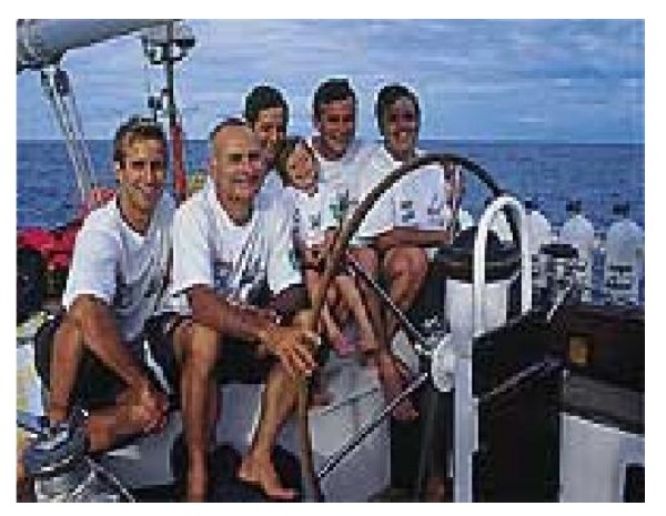 Fotografia de seis pessoas, homens, mulher e crianças, sentadas no convés de um veleiro, junto ao timão, com o mar ao fundo.