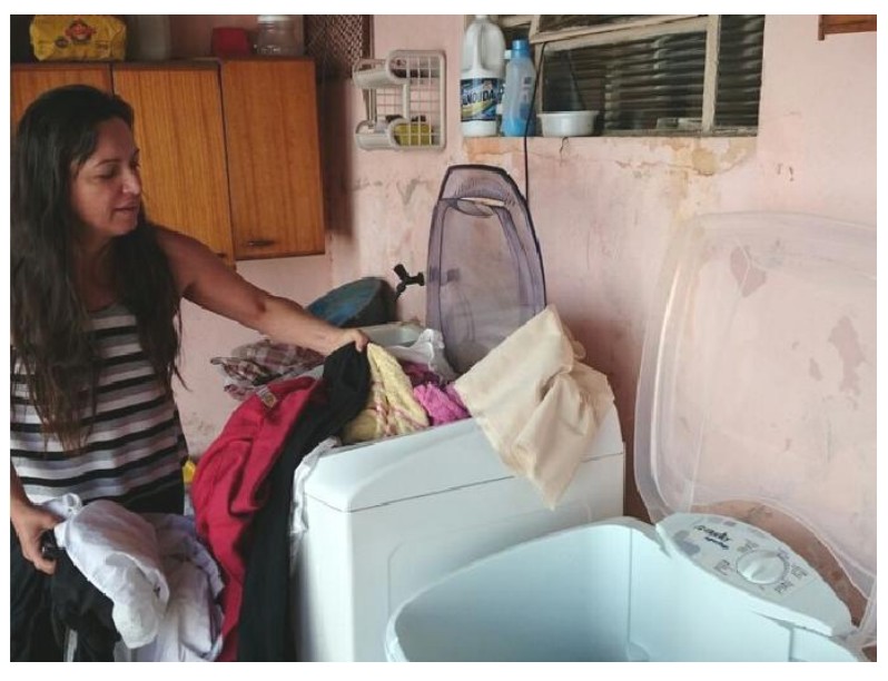 Fotografia de uma mulher colocando roupas em um tanquinho de lavar, em uma área de serviço.