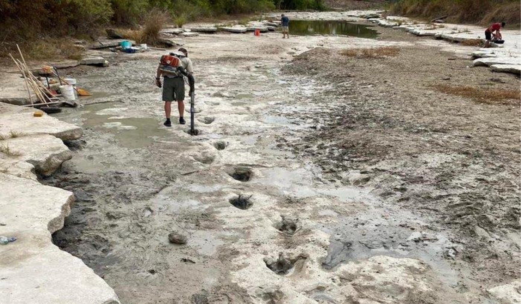 Fotografia do leito seco de um rio, com rochas claras e lama; um homem com equipamento nas costas caminha ao lado de uma sequência de pegadas de três dedos impressas no solo; ao fundo, outras pessoas e uma poça d'água.