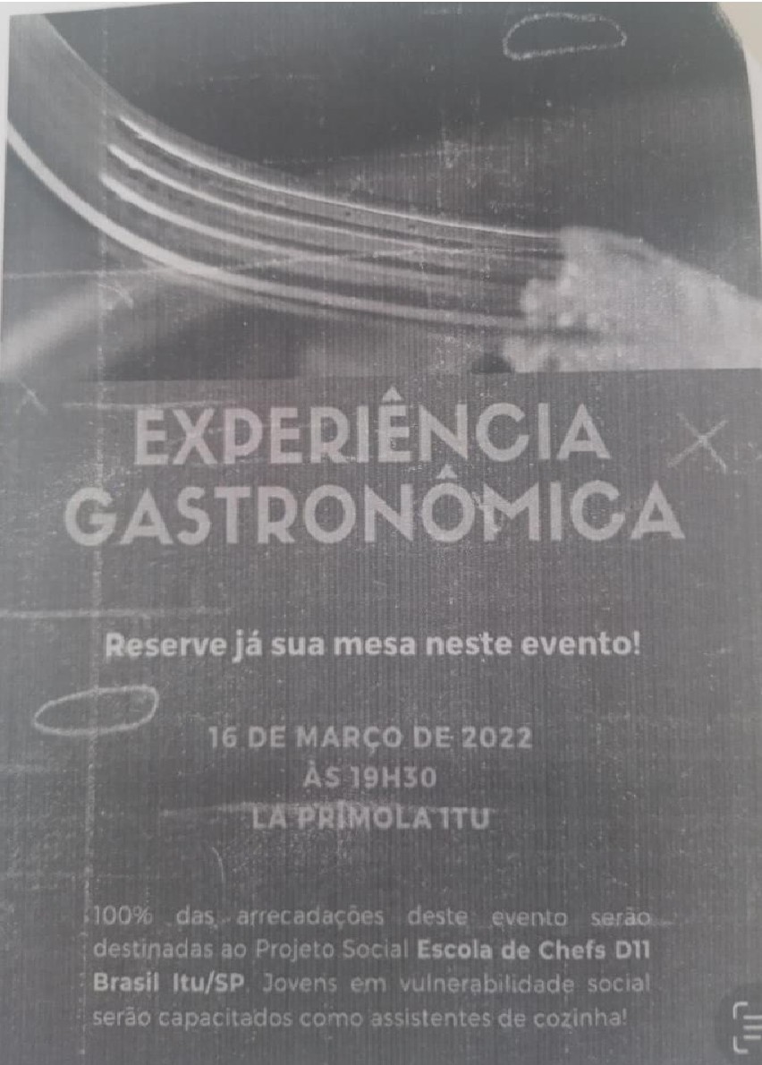 Fotografia de cartaz de evento, em tons de cinza, com garfo estilizado no alto e os dizeres: 'EXPERIÊNCIA GASTRONÔMICA'; 'Reserve já sua mesa neste evento!'; '16 DE MARÇO DE 2022 ÀS 19H30 LA PRÍMOLA ITU'; '100% das arrecadações deste evento serão destinadas ao Projeto Social Escola de Chefs D11 Brasil Itu/SP. Jovens em vulnerabilidade social serão capacitados como assistentes de cozinha!'