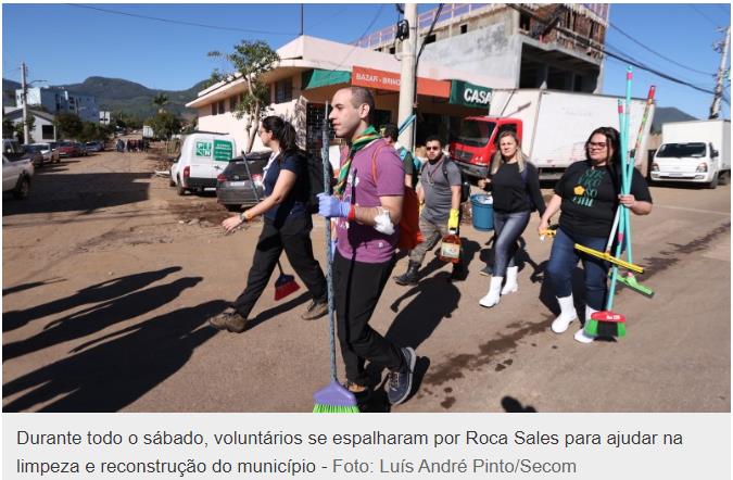 Fotografia: voluntários caminham por uma rua de terra coberta de lama em Roca Sales, carregando vassouras, rodos e baldes; ao fundo, casas, caminhões e um prédio em construção. Legenda impressa na imagem: "Durante todo o sábado, voluntários se espalharam por Roca Sales para ajudar na limpeza e reconstrução do município - Foto: Luís André Pinto/Secom".