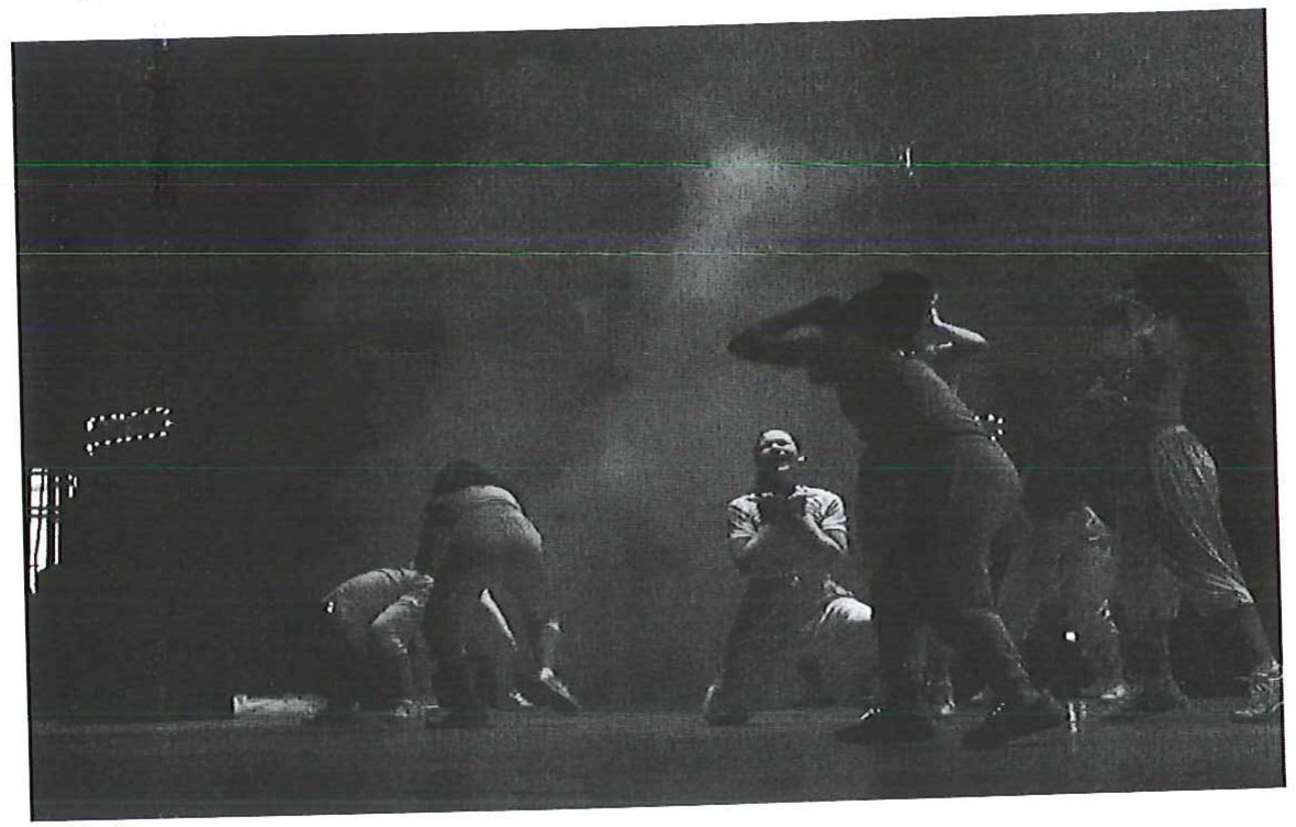 Fotografia em preto e branco de alunos se apresentando em um palco escuro, com fumaça cênica ao fundo.