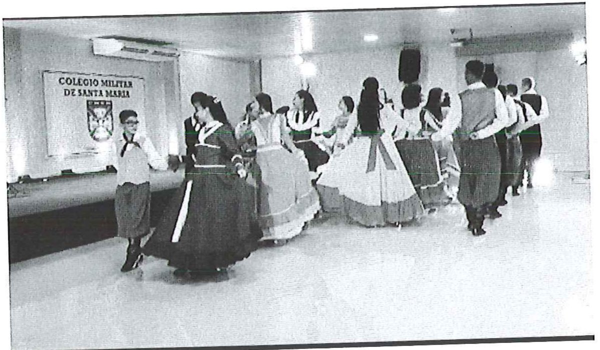 Fotografia de alunos com trajes típicos gaúchos dançando em fila em um salão, com uma placa do Colégio Militar de Santa Maria na parede ao fundo.