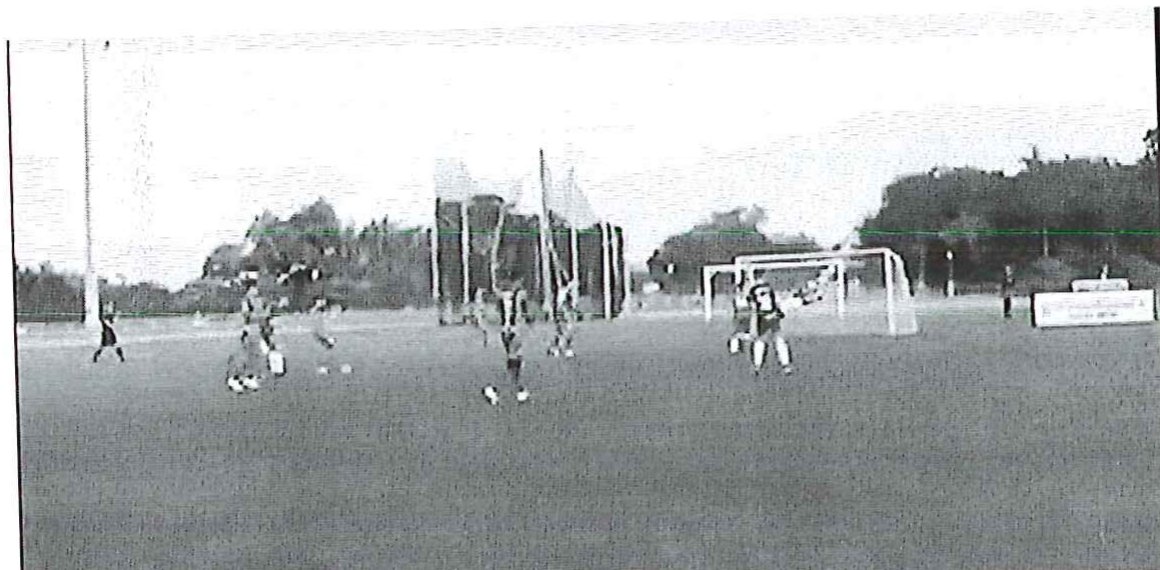 Fotografia de uma partida de futebol society em campo gramado, com jogadores disputando a bola perto de uma das traves.