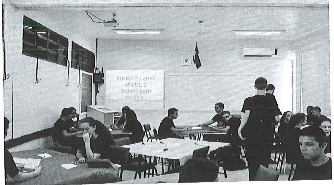 Fotografia de uma sala de aula com alunos sentados em grupos ao redor de mesas, estudando; ao fundo, uma projeção na parede com o texto 'OBMEP / 2018 NÍVEL 2'.