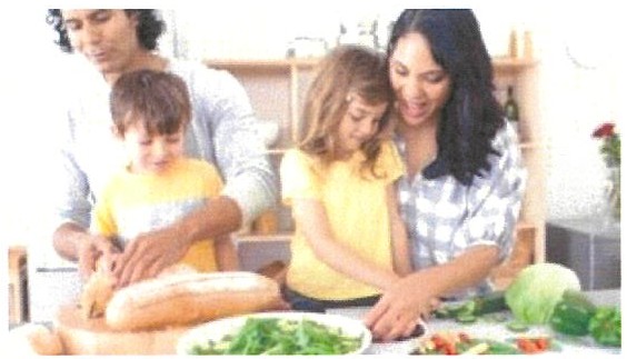 Fotografia: um homem, uma mulher e duas crianças preparam alimentos juntos numa cozinha, cortando pão e verduras sobre a bancada.