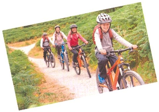 Fotografia: um grupo de crianças pedala em fila numa trilha de terra entre a vegetação verde, usando capacetes.