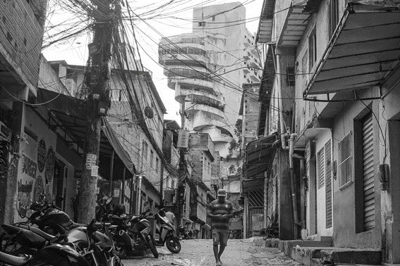 Fotografia em preto e branco de uma viela estreita da favela de Paraisópolis, com casas simples, emaranhado de fios elétricos em postes, motocicletas estacionadas e um homem caminhando; ao fundo, um edifício alto de luxo com varandas.