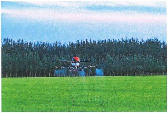 Fotografia de um drone agrícola sobrevoando e pulverizando uma plantação verde, com uma fileira de árvores ao fundo.