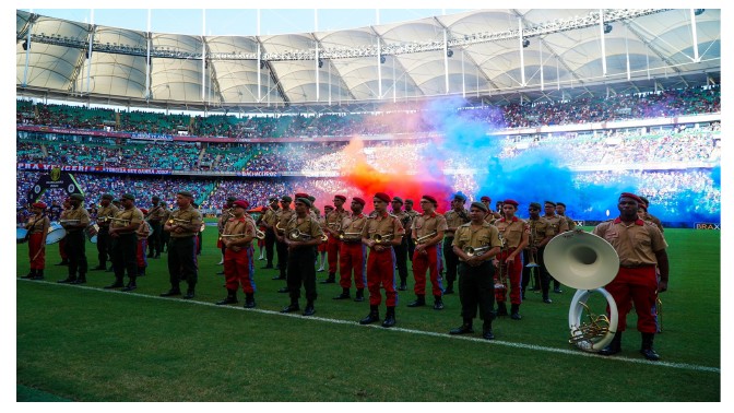 Fotografia da Banda de Música do Exército, em formação, no gramado da Arena Fonte Nova, com fumaça colorida em azul, vermelho e branco ao fundo e arquibancadas com público.
