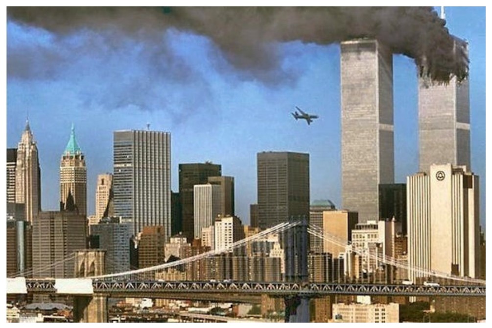 Fotografia do horizonte de Nova York com as torres gêmeas do World Trade Center em chamas, soltando fumaça escura, e um avião se aproximando; em primeiro plano, prédios e uma ponte.