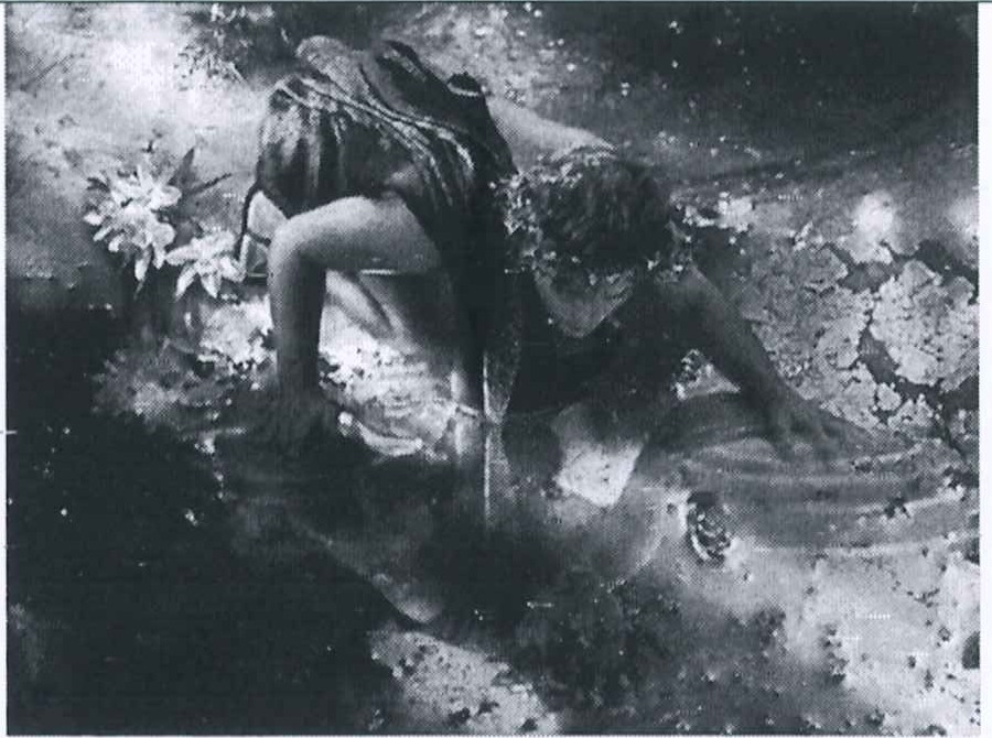 Fotografia em preto e branco de uma pessoa ajoelhada à beira da água, inclinada sobre a superfície como se olhasse o próprio reflexo, com flores ao lado.