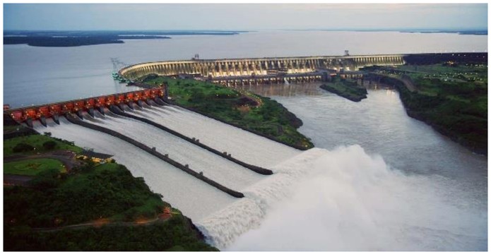 Fotografia aérea de uma grande usina hidrelétrica com barragem curva e vertedouro liberando água para o rio.
