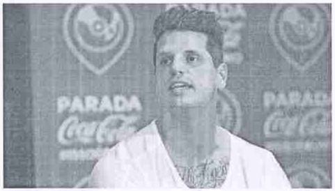 Fotografia de Fernando Fernandes falando, de camiseta branca, diante de painel com os dizeres “Parada Coca-Cola”.