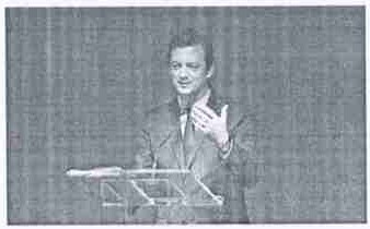 Fotografia de Andrew Parsons, de terno, discursando em um púlpito transparente.