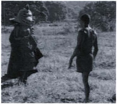 Fotografia de duas figuras em um campo: à esquerda, uma personagem fantasiada de Cuca, com cabeça de jacaré e capa escura; à direita, um menino representando o Saci.