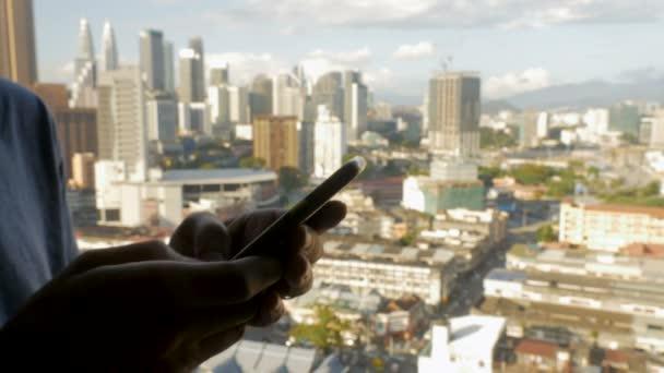 Fotografia: mão segurando um smartphone em primeiro plano, com prédios de uma grande cidade ao fundo.