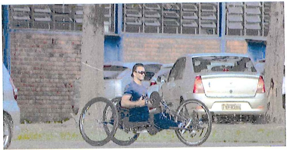 Fotografia de um atleta numa bicicleta adaptada de mão (handbike) em frente a um prédio, com carros estacionados ao fundo.