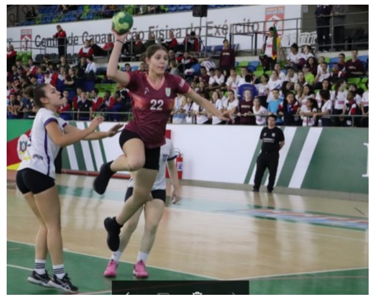 Fotografia de uma partida de handebol feminino em ginásio: atleta de camisa grená número 22 salta com a bola erguida diante de uma adversária de uniforme branco; arquibancada ao fundo.