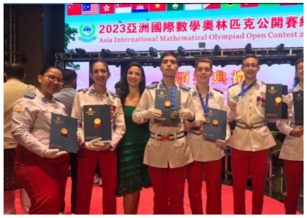 Fotografia de estudantes do CMSM, de uniforme de gala, segurando certificados e medalhas diante de um painel da Asia International Mathematical Olympiad Open Contest 2023.