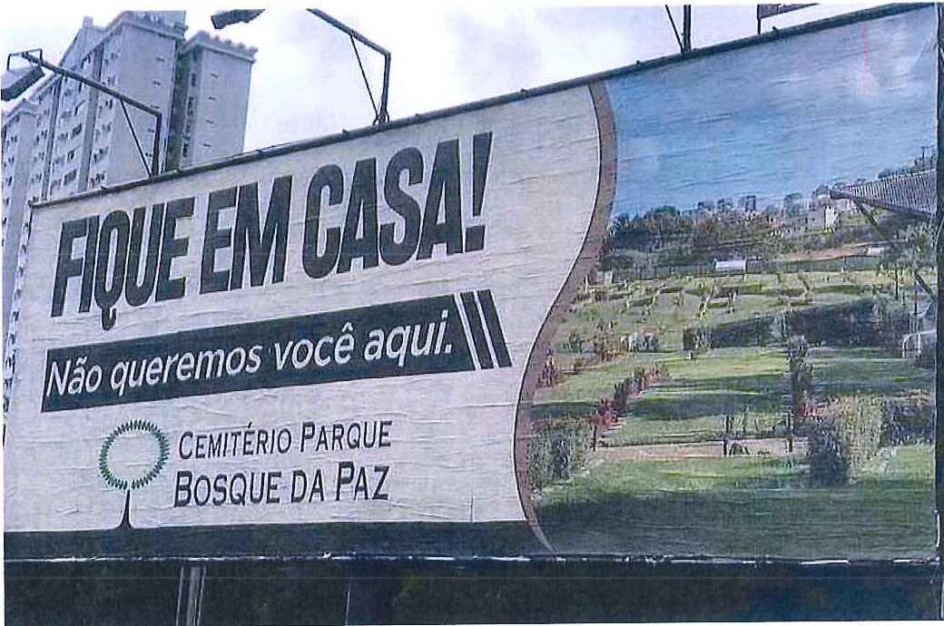 Fotografia de um outdoor com os dizeres "Fique em casa! Não queremos você aqui." e a marca "Cemitério Parque Bosque da Paz", ao lado de uma foto de um cemitério-parque gramado.