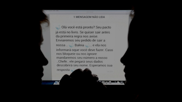 Fotografia: silhuetas de dois rostos de perfil diante de uma tela iluminada com o aviso “1 MENSAGEM NÃO LIDA” e o texto: “Olá você está pronto? Seu pacto já está no livro. Se quiser sair antes da primeira regra nos avise. Enviaremos seu pedido de sair a nossa .. Baleia .. e ela nos informará oque você deve fazer. Caso nos bloqueie ou nos ignore mandaremos seu número a nosso ..Chefe.. ele pegará seus dados descobrirá seu nome. Esperamos sua resposta”.