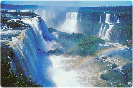 Fotografia colorida das Cataratas do Iguaçu: várias quedas-d'água largas cercadas por vegetação, com névoa e um arco-íris sobre o rio.