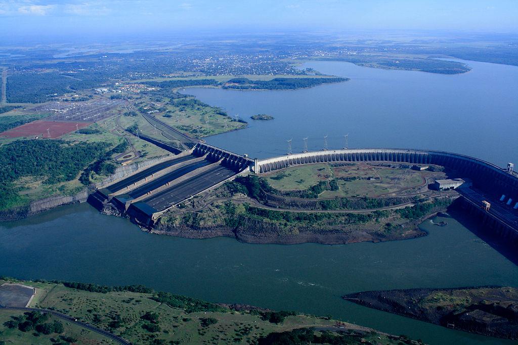 Fotografia aérea colorida da Usina Hidrelétrica de Itaipu: a barragem atravessando o rio, com o vertedouro à esquerda e o grande lago da represa ao fundo.