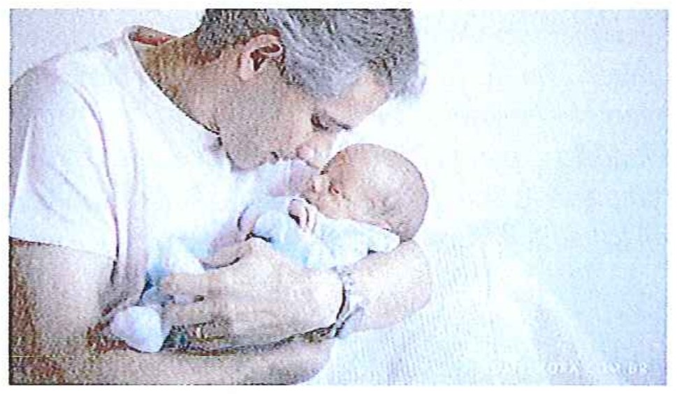 Fotografia de um homem de cabelos grisalhos, de camiseta branca, beijando a cabeça de um bebê recém-nascido que segura no colo.