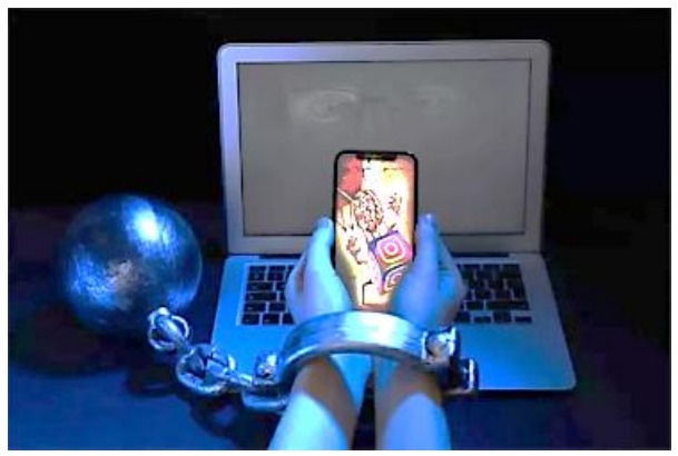Fotografia de mãos algemadas segurando um celular diante de um notebook; a algema está presa por corrente a uma bola de ferro.