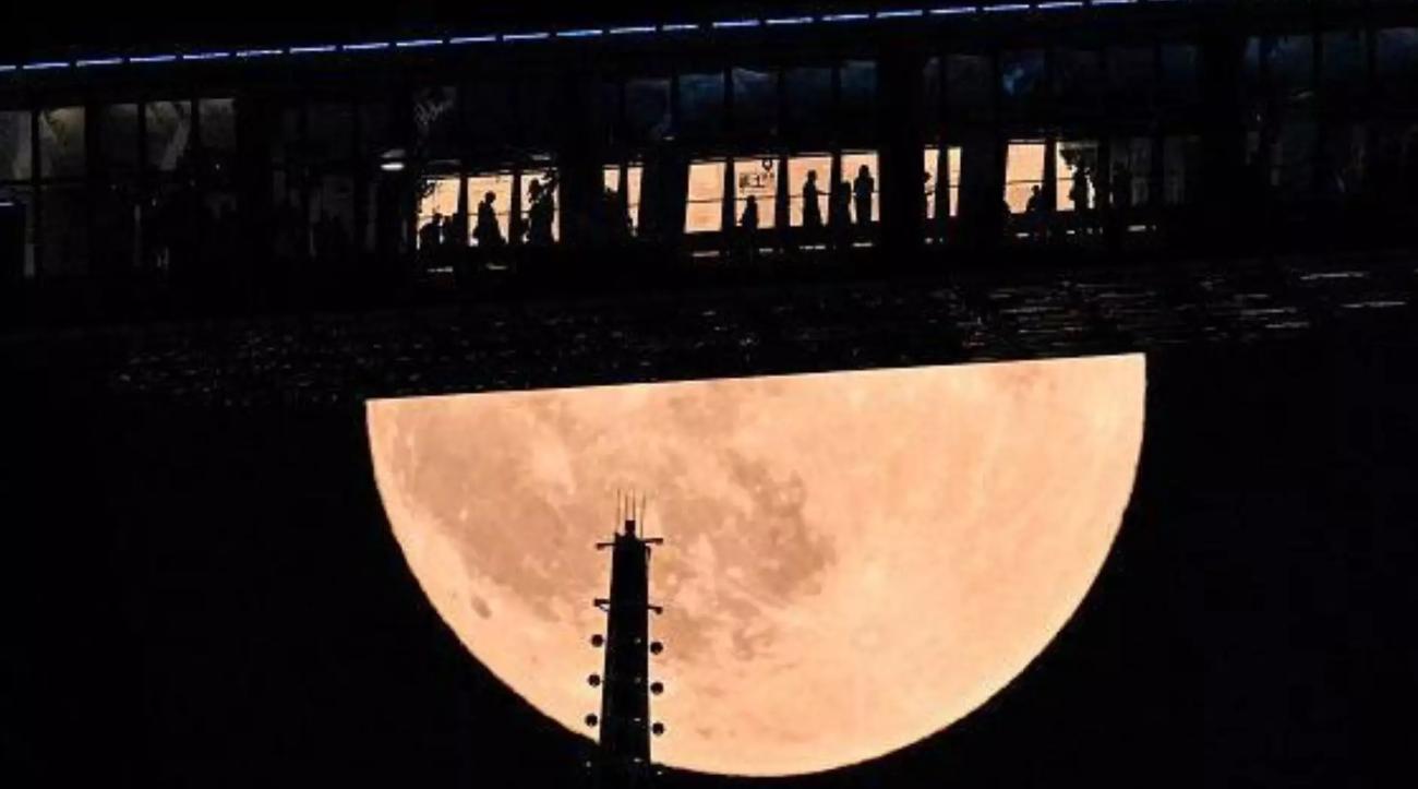 Fotografia noturna da Lua cheia, de cor alaranjada, parcialmente encoberta por uma passarela escura onde se veem silhuetas de pessoas; à frente da Lua aparece uma torre estreita com pequenas luzes.