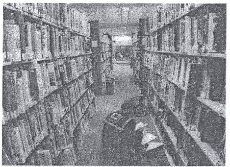Fotografia em preto e branco de um corredor estreito de biblioteca entre estantes altas repletas de livros, com uma pessoa sentada no chão ao fundo.