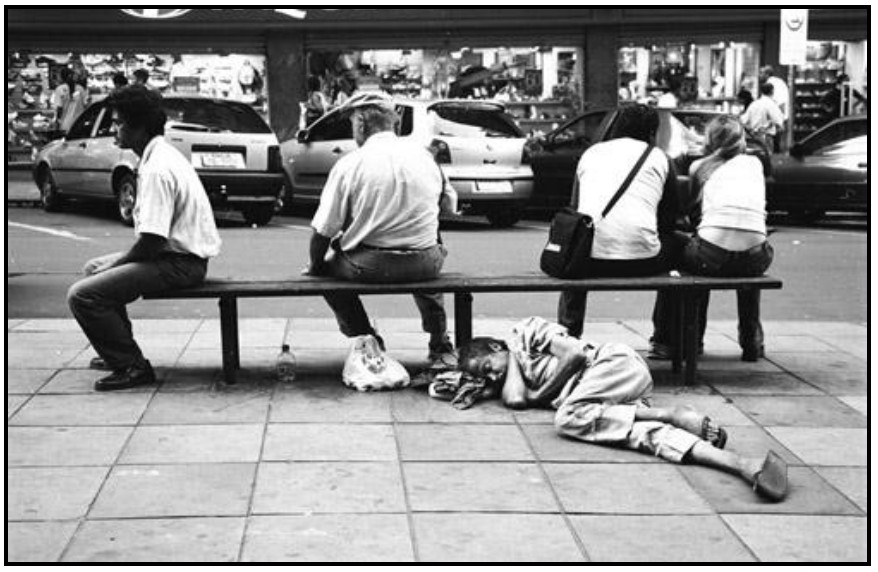 Fotografia em preto e branco de uma rua de cidade: quatro pessoas sentadas de costas em um banco de praça, e um homem maltrapilho deitado no chão, debaixo do banco, aparentemente dormindo.