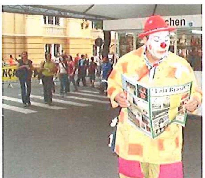 Fotografia colorida de um palhaço com chapéu vermelho e roupa colorida lendo um jornal em uma rua movimentada.