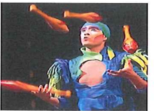 Fotografia de malabarista com roupa azul e verde lançando claves no ar.