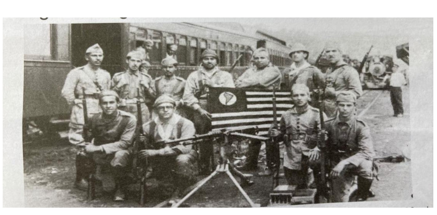 Fotografia em preto e branco de soldados uniformizados posando junto a um trem, com metralhadora no chão e a bandeira paulista ao centro.