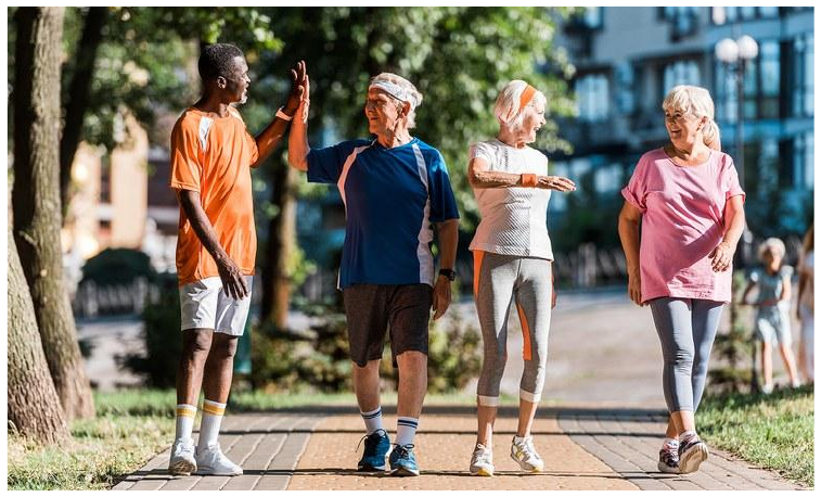 Fotografia de quatro pessoas caminhando sorridentes em um parque arborizado: um jovem de camiseta laranja cumprimenta, com as mãos erguidas, um homem idoso de faixa na cabeça; ao lado, duas mulheres idosas em roupas esportivas.