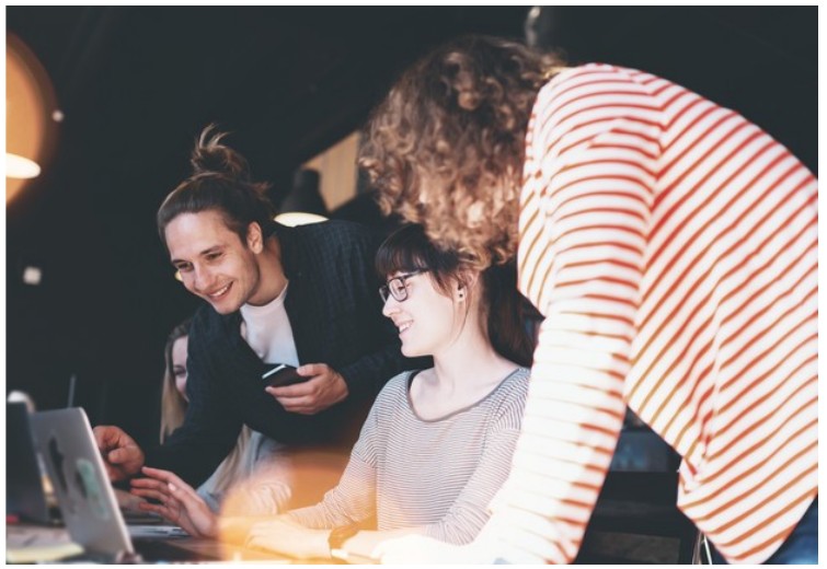 Fotografia de jovens sorrindo diante de um notebook; uma pessoa usa um celular.