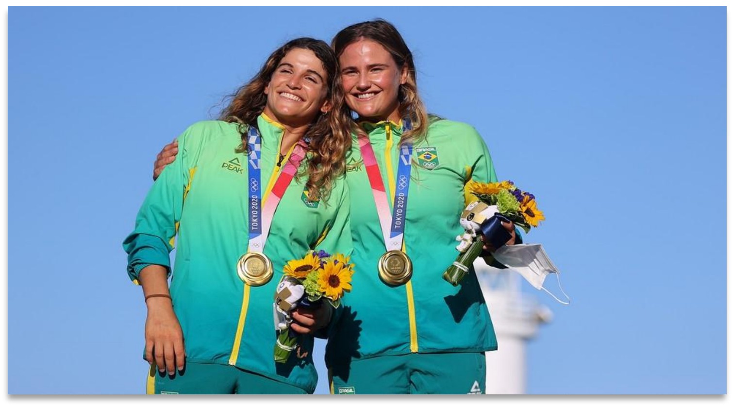 Fotografia de duas velejadoras sorridentes, abraçadas, com uniformes verde e amarelo do Brasil, medalhas de ouro no pescoço e buquês de flores nas mãos, sob céu azul.