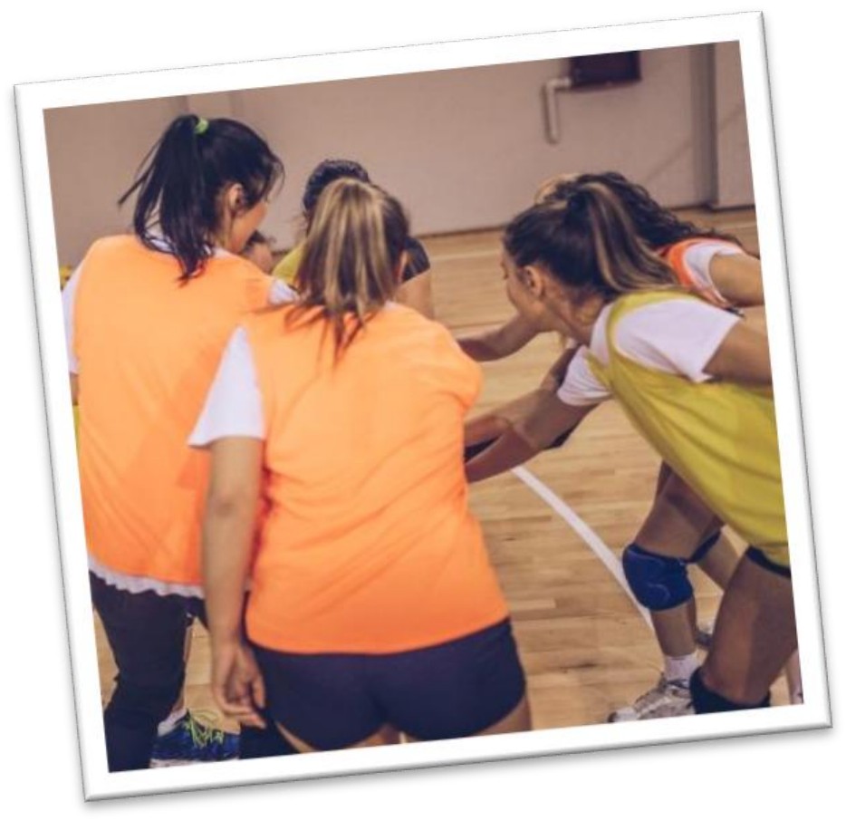 Fotografia de um grupo de jogadoras com coletes laranja e amarelos reunidas em círculo, abraçadas, numa quadra esportiva.