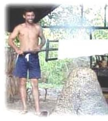 Fotografia de um seringueiro sorridente, de bermuda, ao lado de um grande bloco de borracha defumada.