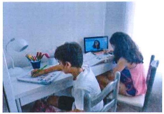 Fotografia de duas crianças estudando em uma escrivaninha em casa, com um computador mostrando uma videochamada.