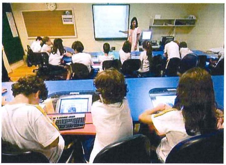 Fotografia de uma sala de aula com alunos usando notebooks em bancadas e uma professora diante de um quadro de projeção.
