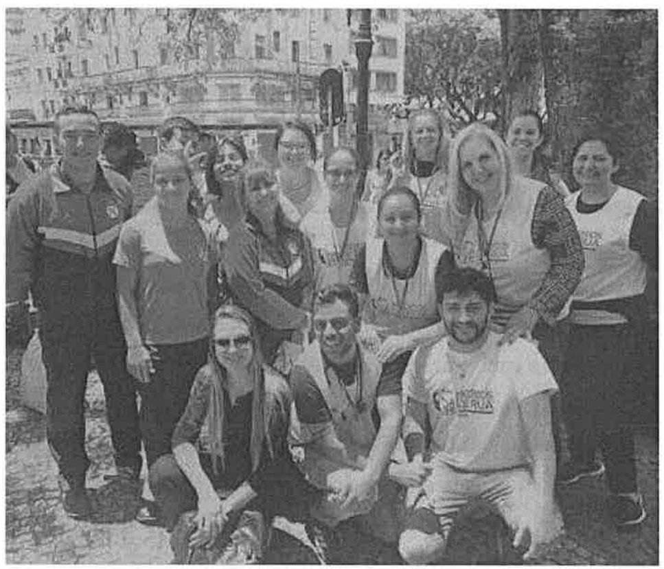 Fotografia de um grupo de cerca de quinze pessoas reunidas ao ar livre em uma praça, posando para a foto; várias vestem coletes claros e há prédios e árvores ao fundo.