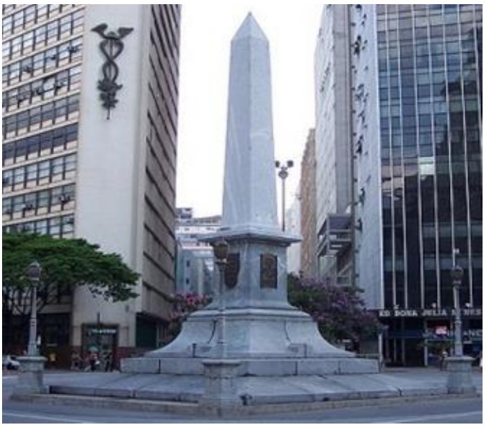 Fotografia do Pirulito da Praça Sete: obelisco de pedra clara sobre pedestal escalonado, com placas na base, cercado por prédios.